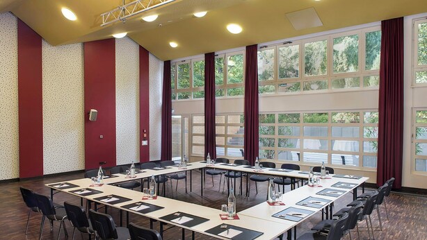 Een vergaderzaal met een lange tafel, stoelen en grote ramen voorzien van gordijnen.