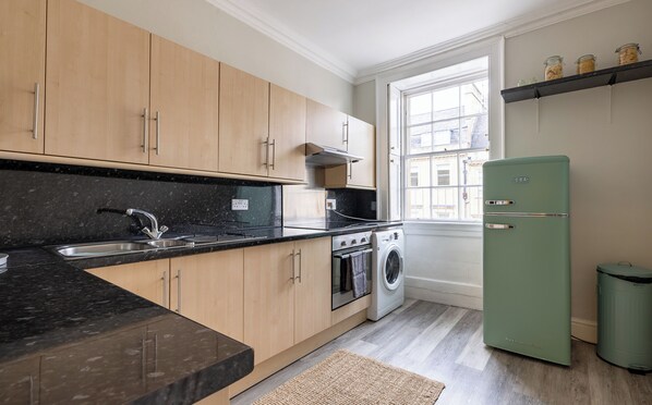 Een moderne keuken met houten kastjes, een zwart aanrecht, een roestvrijstalen spoelbak, een inbouwoven, een wasmachine en een groene koelkast.