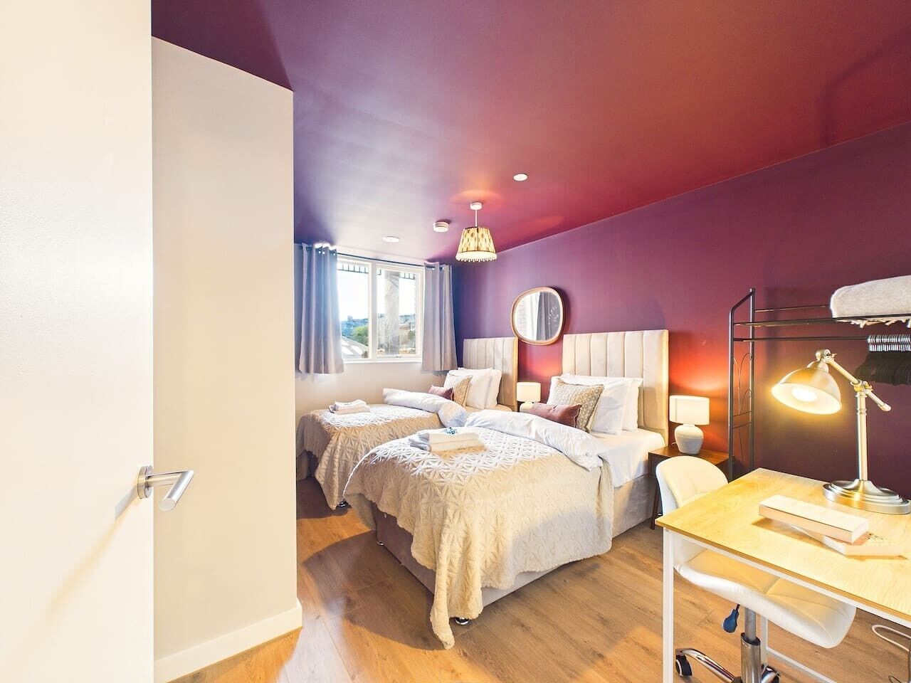 A hotel room with two beds, a desk with a lamp, and a view of the city through the window.