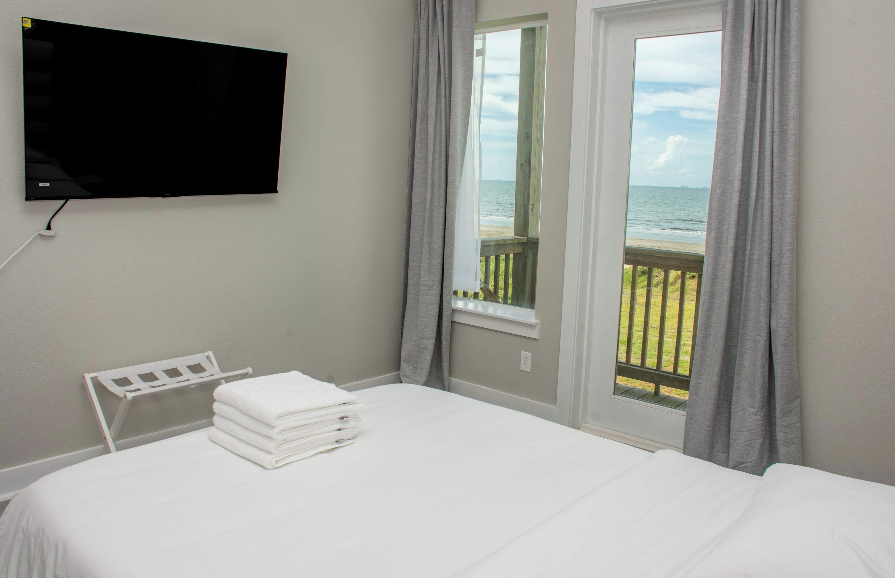 A hotel room with a bed, a TV mounted on the wall, a window with a view of the beach, and a window with curtains.