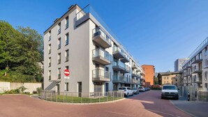 Un edificio moderno de apartamentos con varios balcones, una señal de 'prohibido el paso' y coches estacionados en una calle adoquinada de ladrillo.