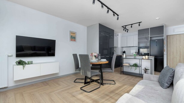 Una sala moderna con televisor de pantalla plana, sofá gris, comedor con sillas y cocina con refrigerador y horno.