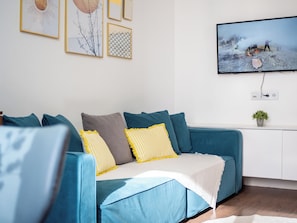 A modern living room with a blue sofa, yellow and grey cushions, a white throw blanket, and a flat-screen TV mounted on the wall.