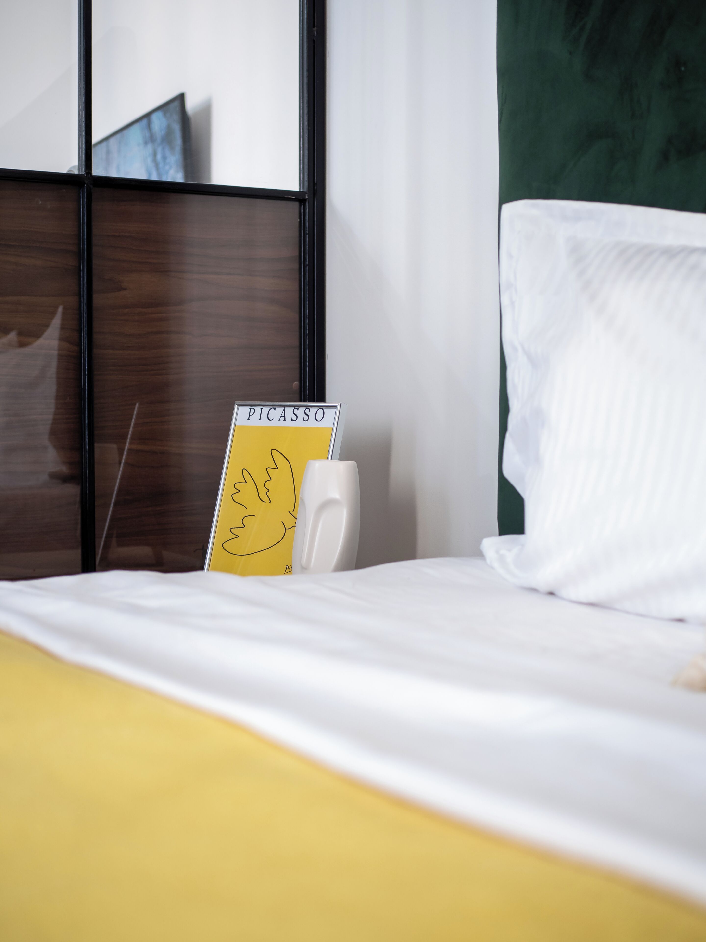 A hotel room with a bed, a bedside table, a white pillow, a yellow sign with the word 'PICASSO' and a drawing, and a green wall.