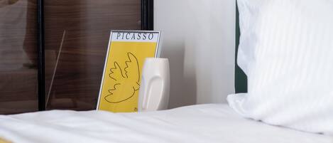 A hotel room with a bed, a bedside table, a white pillow, a yellow sign with the word 'PICASSO' and a drawing, and a green wall.
