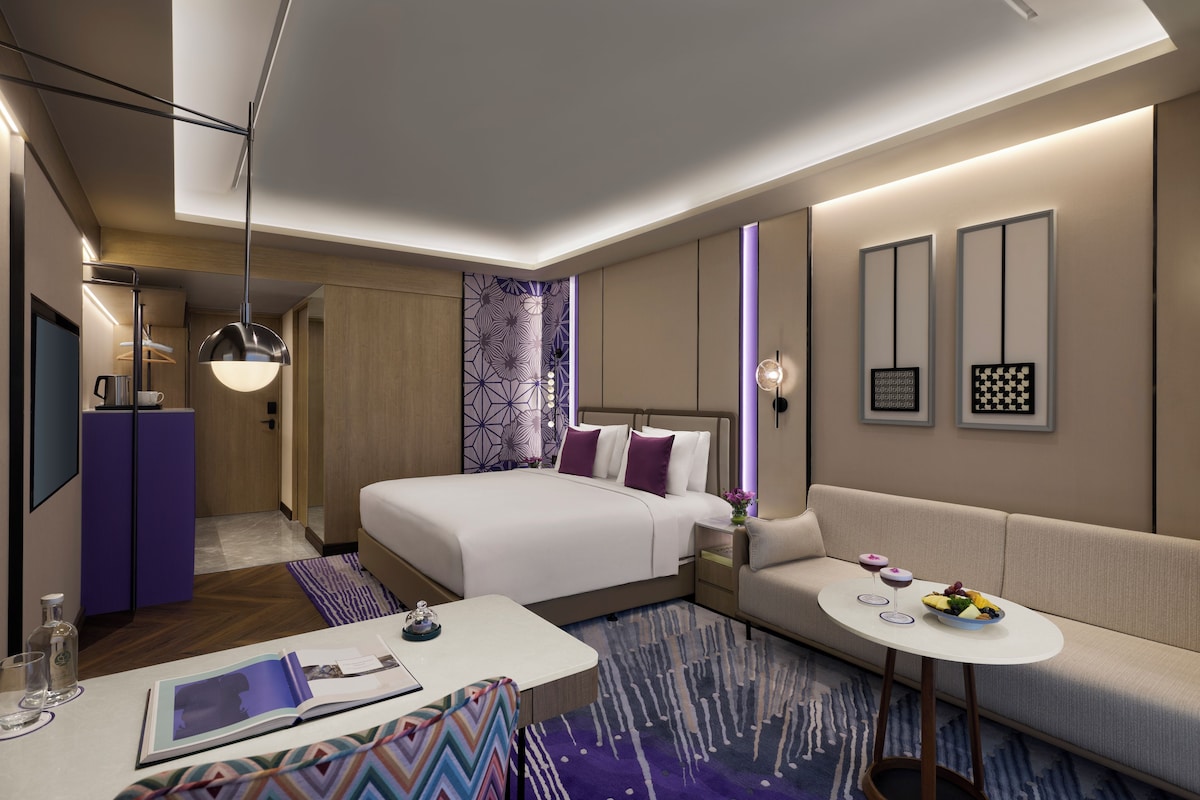 A modern hotel room with a bed, a sofa, a small table with a magazine, and a TV.