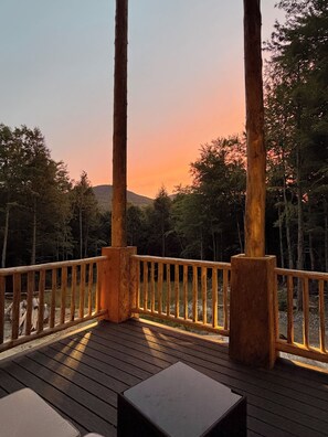 Una terraza de madera con barandilla, una mesa y vistas a un bosque y al atardecer.