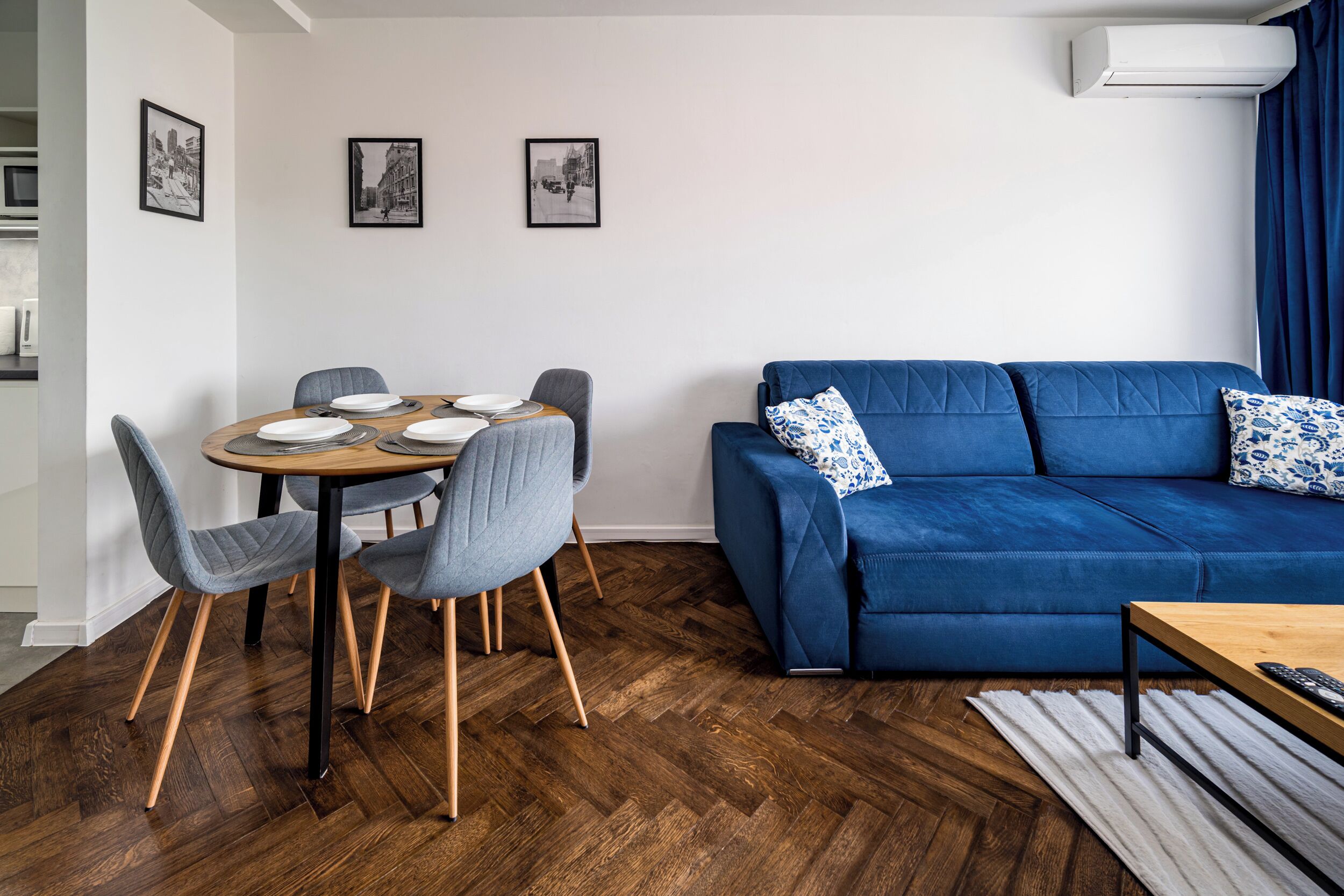A modern living room with a blue sofa, grey chairs, a wooden dining table set for four, and a herringbone wood floor.