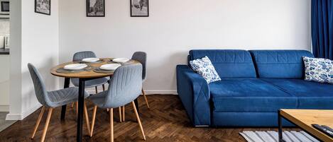 A modern living room with a blue sofa, grey chairs, a wooden dining table set for four, and a herringbone wood floor.