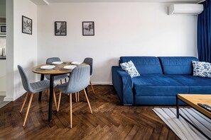 Uma sala de estar moderna com um sofá azul, cadeiras cinzas, uma mesa de jantar de madeira para quatro pessoas e um piso de madeira em padrão herringbone.