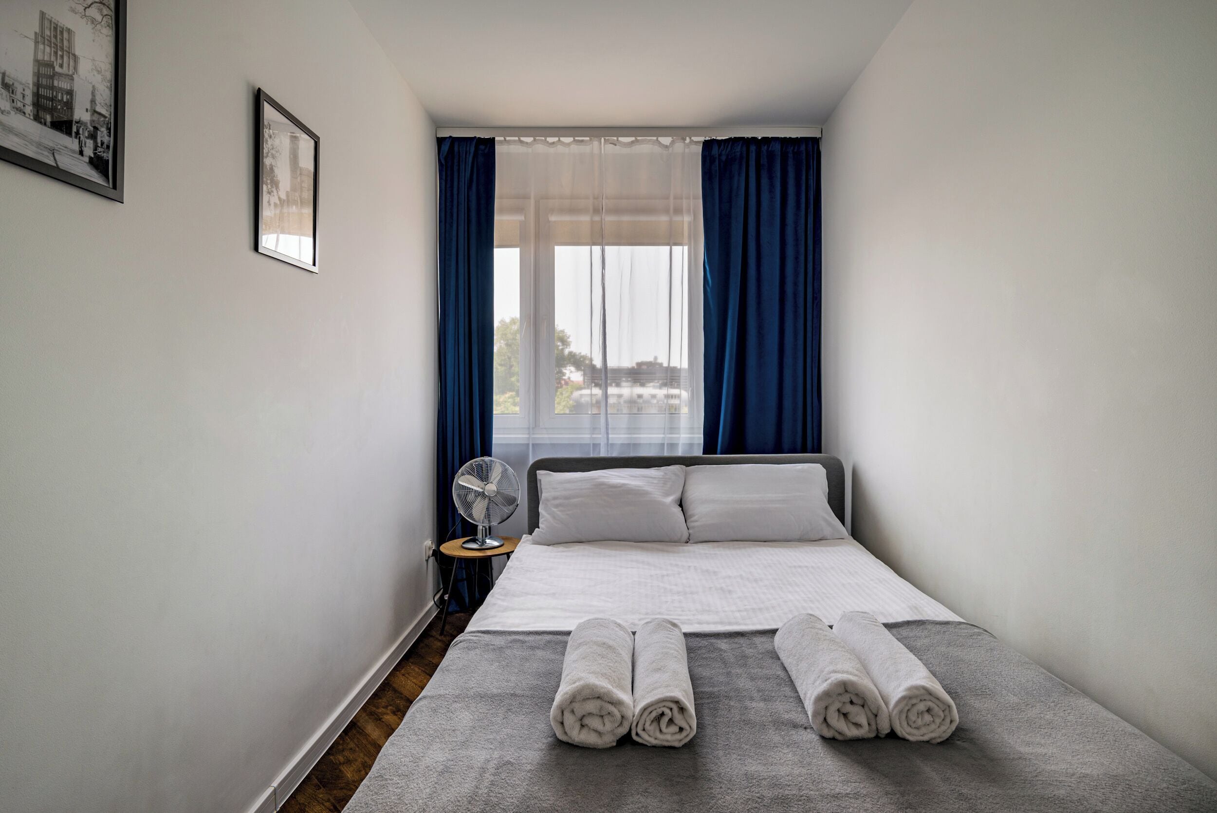 Een compacte, verzorgde hotelkamer met een bed, een nachtkastje met een ventilator en een raam met gordijnen.