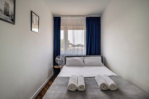 Quarto de hotel pequeno e bem organizado, com cama, mesa de cabeceira com um ventilador e janela com cortinas.