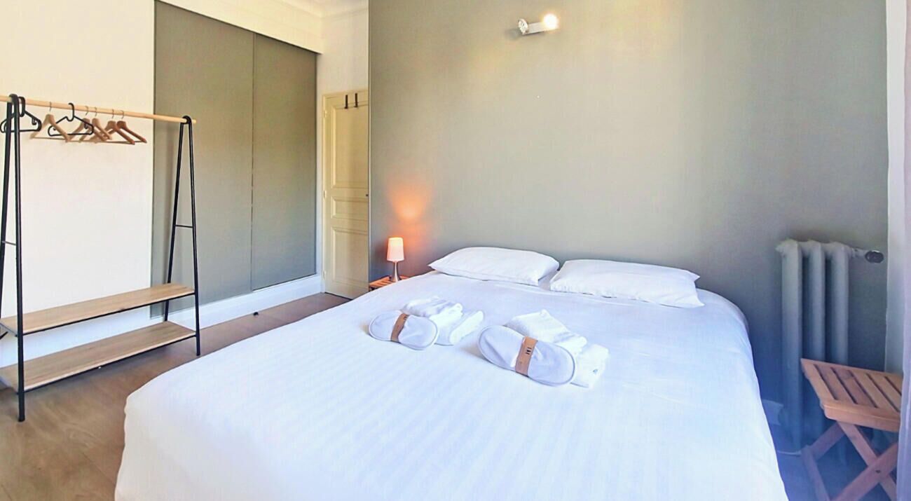 A neatly made bed with white linens and towels, a wooden bench, a wall-mounted light, and a radiator.