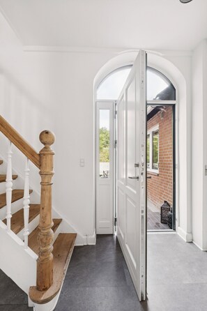 A hallway with a wooden staircase, a white door, and a view of a brick building outside.