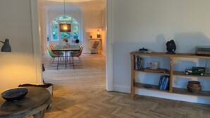 Huoneessa on kalanruotokuviollinen lattia, ruokailutila, jossa on pöytä ja tuolit, kirjahylly kirjoineen ja koriste-esineineen sekä ikkuna, josta avautuu näkymä vehreään maisemaan.