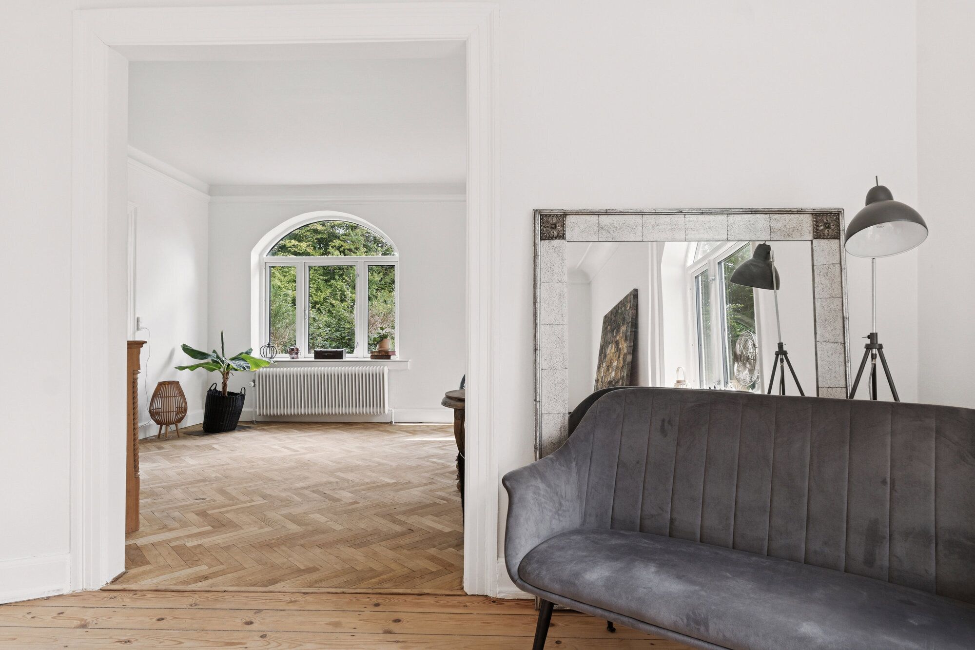 A room with a grey sofa, a large mirror, and a window with a view of greenery.