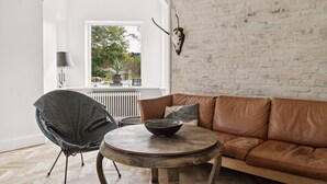 A room with a brick wall, a brown leather sofa, a round wooden table with a bowl, a woven chair, and a window with a view of greenery. - Slap af I Nordsjælland i dette hyggelige hus med en dejlig have. (Kvistgård)