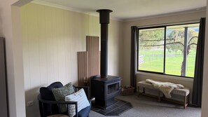 Una sala de estar con un estufa de leña negra, un sofá con cojines, un banco y vistas a un paisaje verde a través de un gran ventanal.