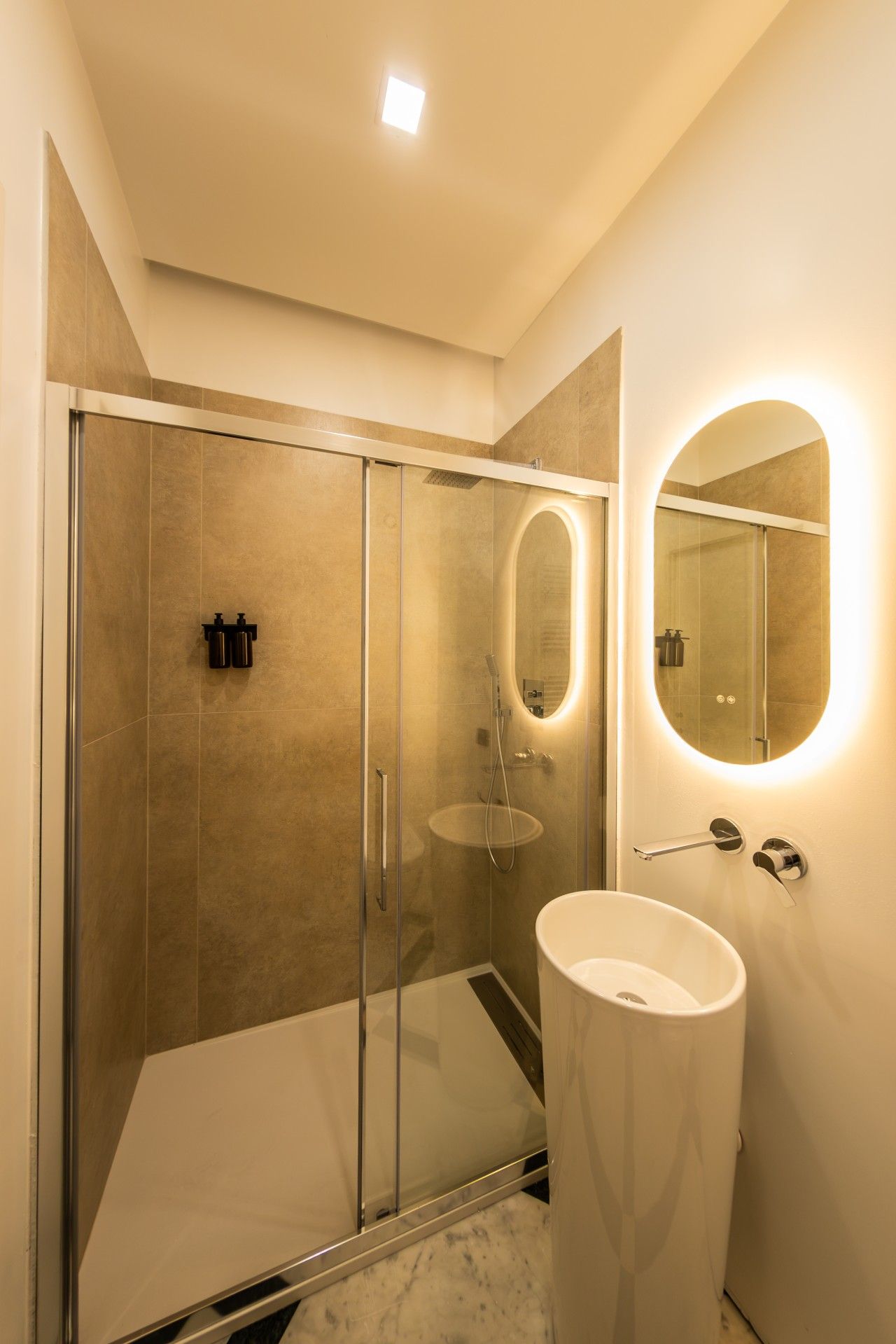 Une salle de bain moderne avec une douche à parois de verre, un grand miroir rond avec éclairage intégré et un lavabo blanc.