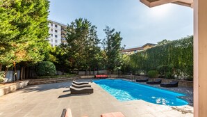 Ein Schwimmbecken mit Liegestühlen und einer Steindeck, umgeben von Bäumen und einem Gebäude im Hintergrund.
