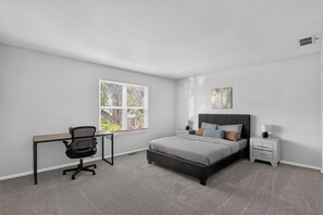 A bedroom with a bed, a desk, a chair, a bedside table, and a window with blinds. - Comfortable Townhome with Modern Interiors (Indianapolis)