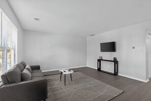 Uma sala de estar moderna com um sofá cinzento, uma mesa de centro, uma televisão na parede e um tapete estampado.