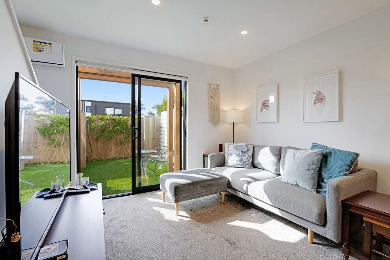 A modern living room with a grey sofa, a coffee table, and a TV. There is a sliding glass door leading to a backyard.