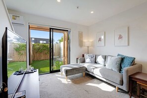 A modern living room with a grey sofa, a coffee table, and a TV. There is a sliding glass door leading to a backyard.