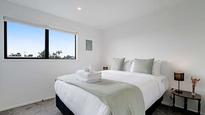 A modern hotel room with a large bed, a bedside table with a lamp, and a view through a window.