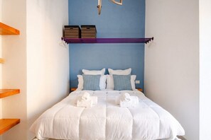 A neatly made bed with white bedding and two pillows, a wooden shelf to the left, and a blue wall with a light fixture above.