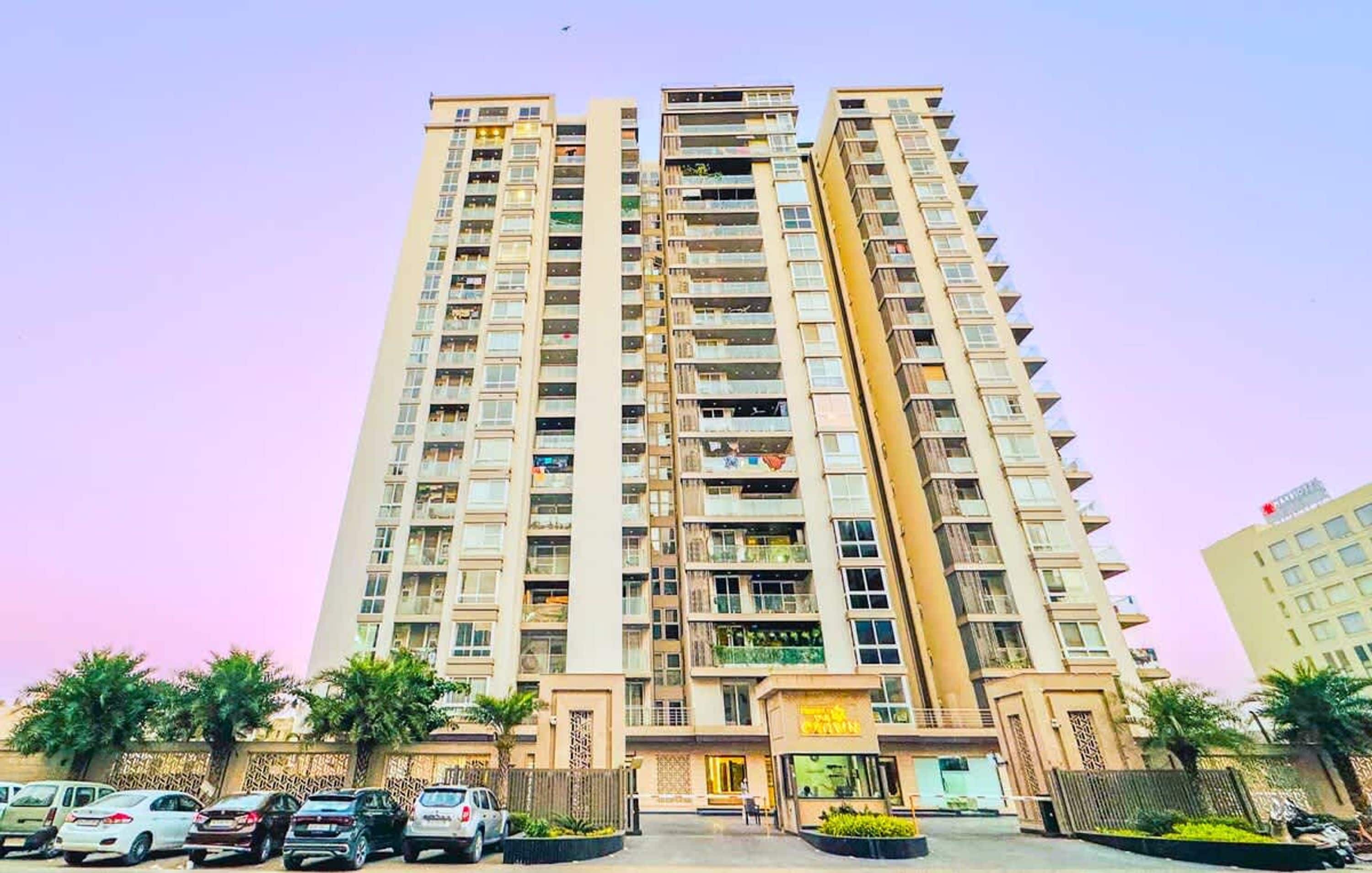 Un edificio residencial alto con varios balcones, entrada con acceso restringido y un estacionamiento con automóviles.