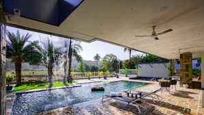 Ein Poolbereich mit Blick auf Palmen und eine weite Landschaft, ein überdachter Sitzbereich mit Liegestühlen und einem Deckenventilator.