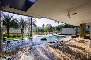 A pool area with a view of palm trees and a distant landscape, a covered seating area with lounge chairs, and a ceiling fan.