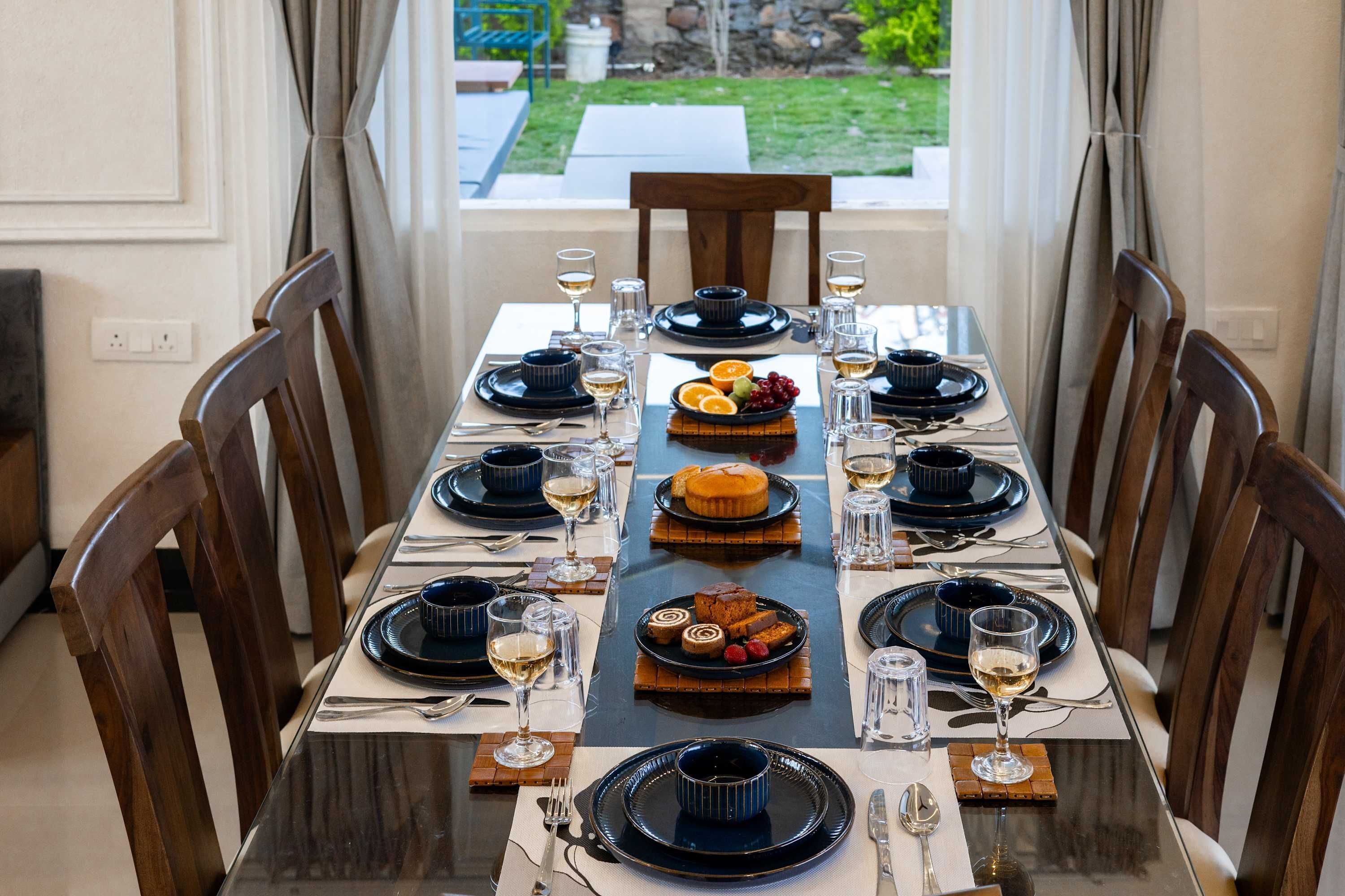 Mesa de jantar posta para seis, com pratos pretos, copos transparentes e diversas sobremesas.