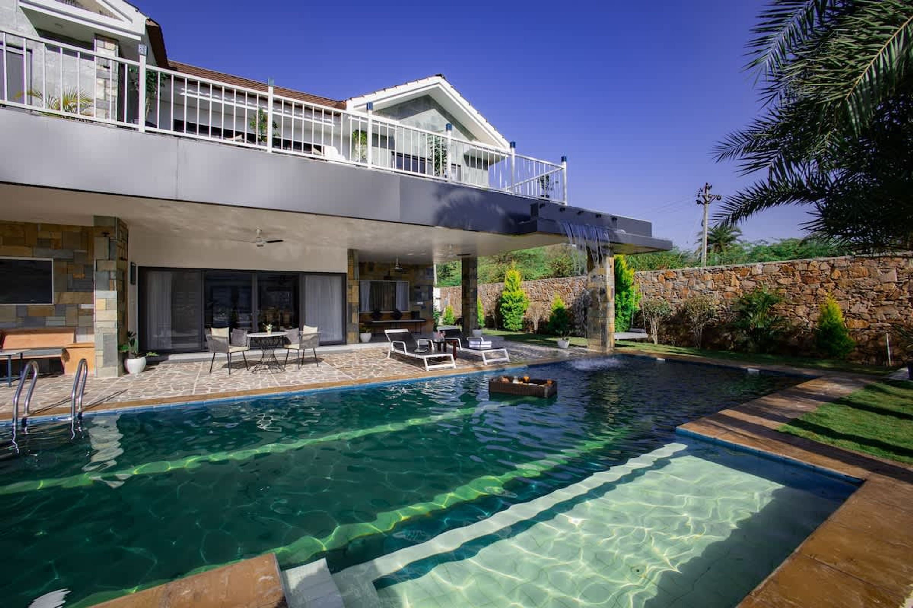 A modern two-story house with a swimming pool, outdoor seating, and a stone wall.