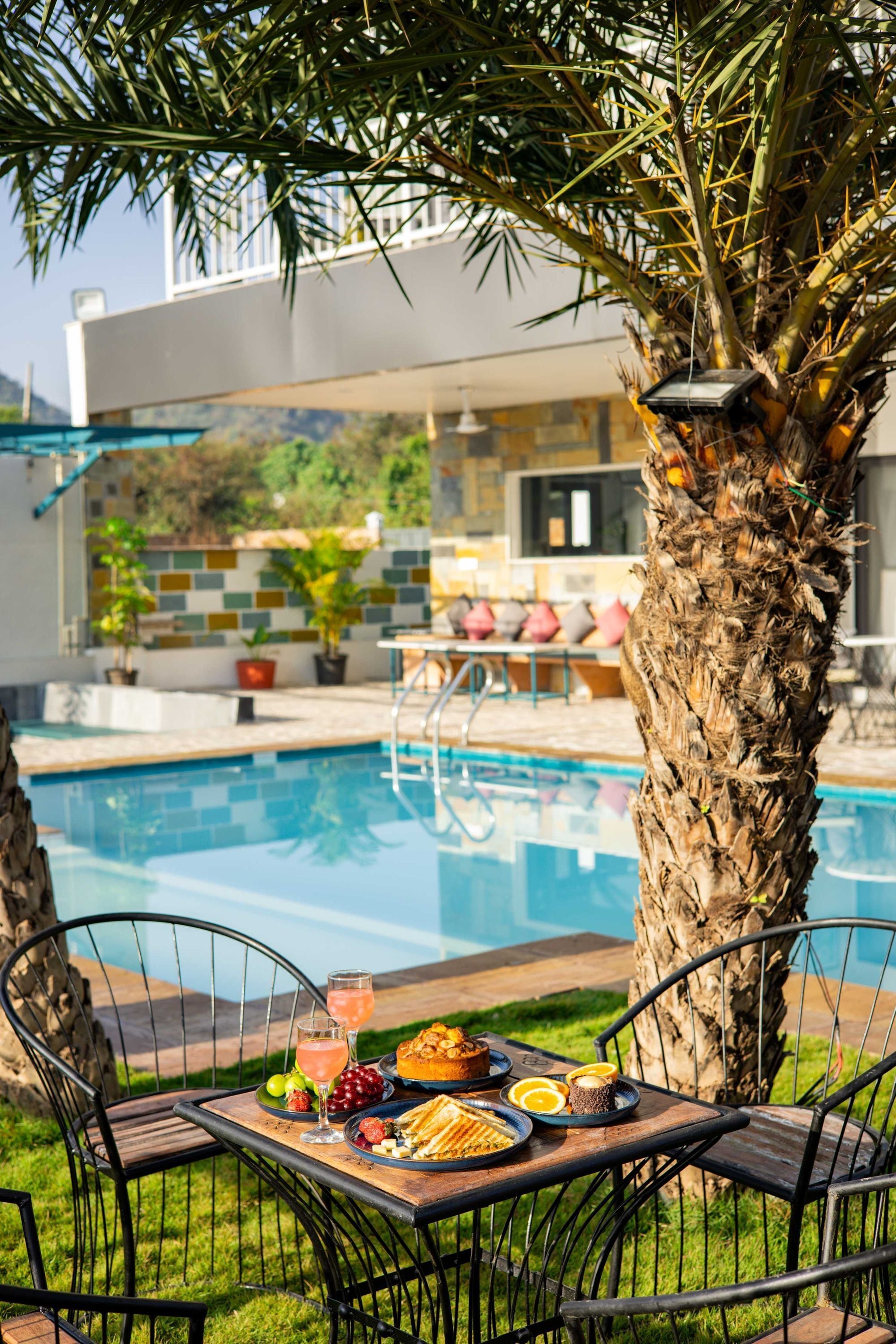 Area pranzo a bordo piscina con tavolo apparecchiato per due, una piscina e un edificio moderno sullo sfondo.