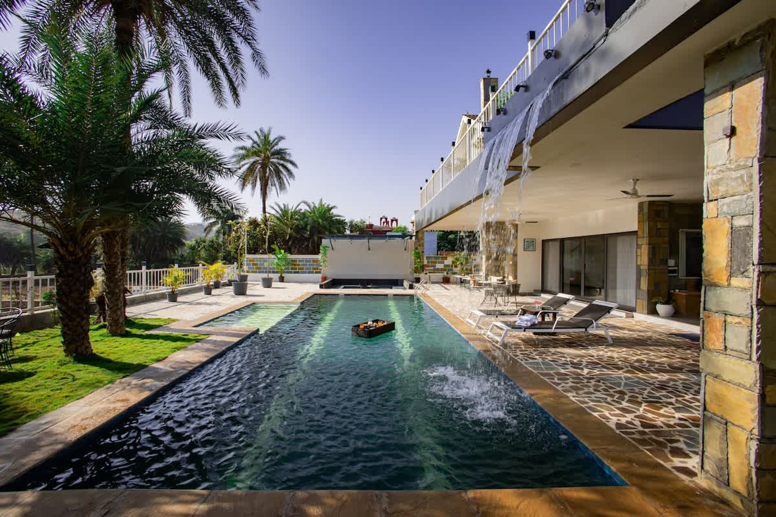 A swimming pool with a water feature, surrounded by palm trees and a stone-tiled deck.