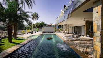 A swimming pool with a water feature, surrounded by palm trees and a stone-tiled deck.