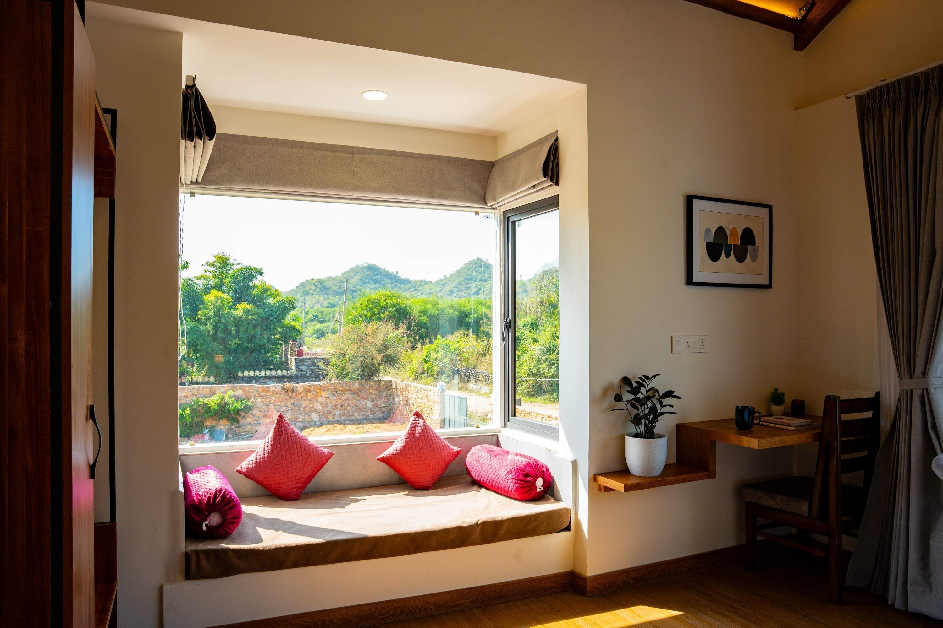 A room with a large window, a wooden bench with red cushions, a small table with a plant, and a framed picture on the wall.
