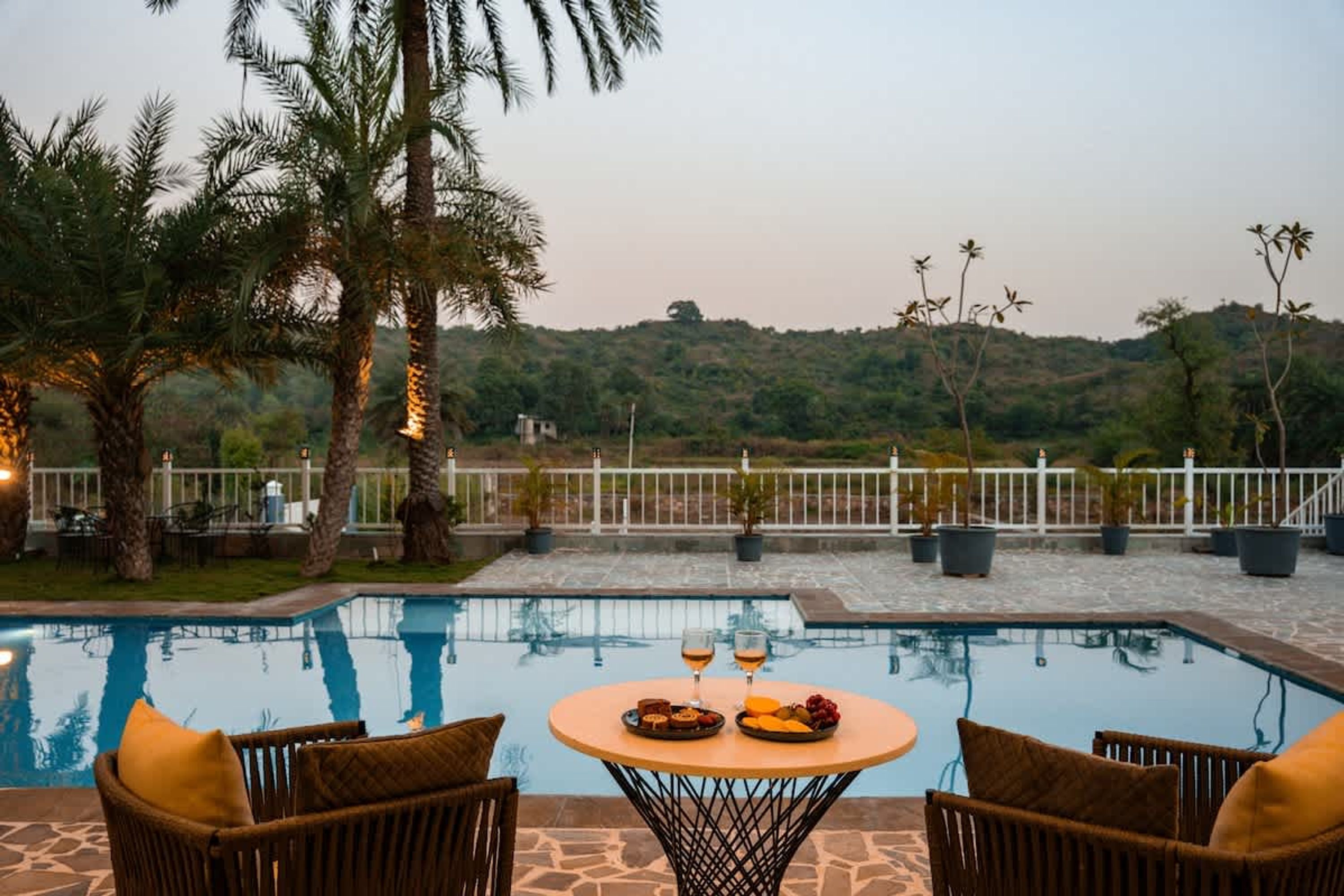 An outdoor pool area with a table set for two, surrounded by chairs, and overlooking a hilly landscape.