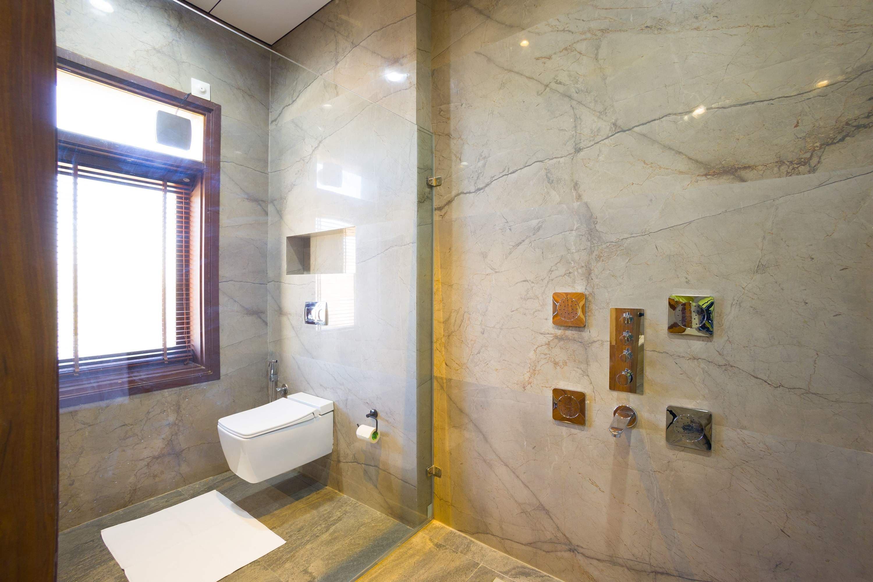 A modern bathroom with a marble wall, a toilet, a shower, and a window with blinds.