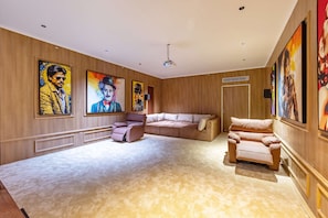 A spacious living room with a large sectional sofa, a recliner, and a coffee table. The walls are adorned with large, colorful paintings. - Kvanya Mansion by Viraalay - Luxe 5bhk Villa in Udaipur (Udaipur)