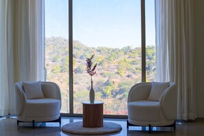 Ein Zimmer mit zwei Stühlen, die zu einem großen Fenster zeigen, einer runden Tischplatte mit einer Pflanze in einer Vase und Blick auf eine hügelige Landschaft.