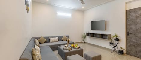 Uma sala de estar moderna com um sofá seccional cinza, uma mesa de centro e uma TV de tela plana na parede. Há plantas em vasos sobre um suporte de metal preto e um ventilador de teto.
