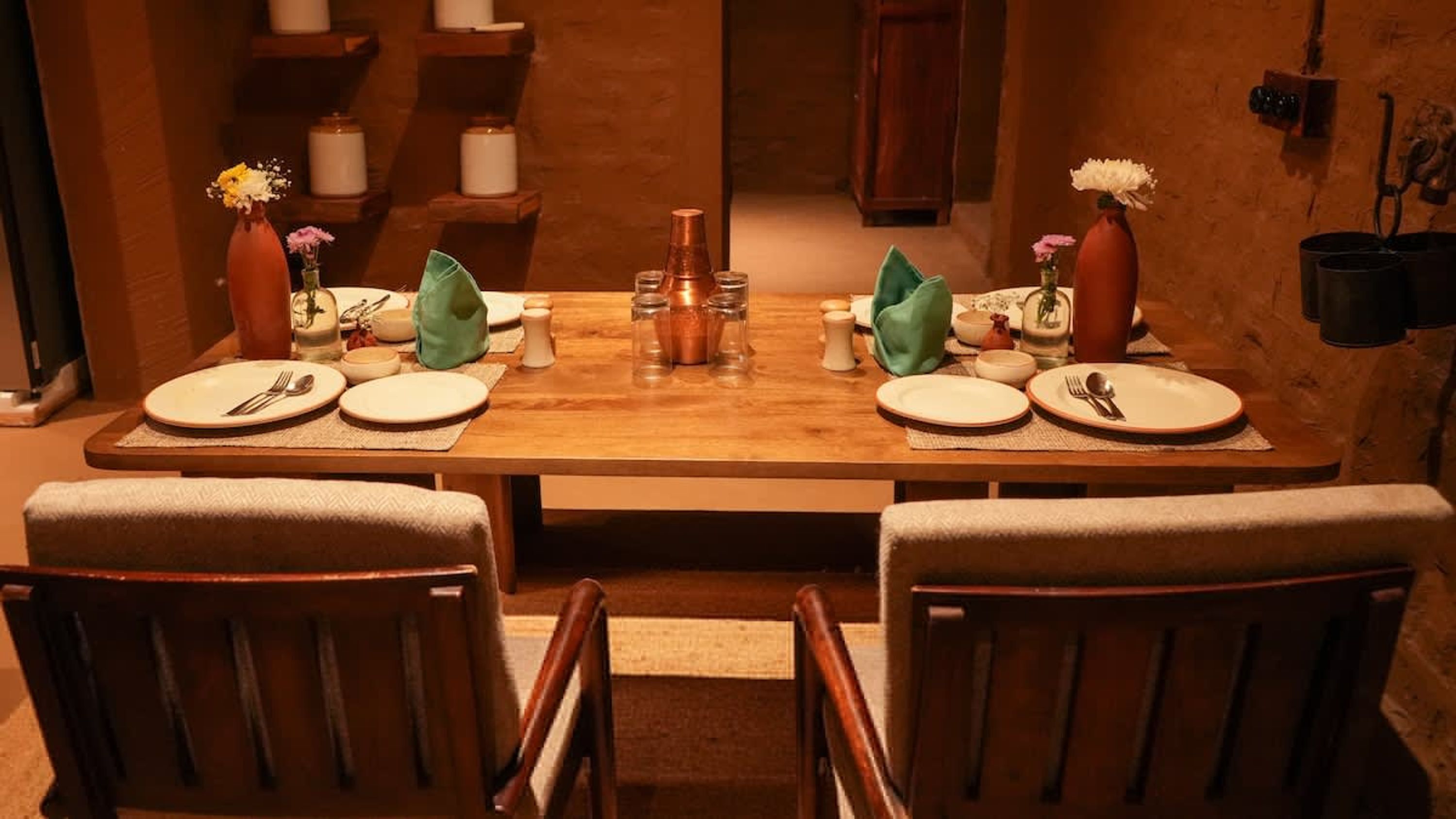 Mesa de comedor preparada para dos, con sillas, un jarrón de flores, saleros y pimenteros, y una olla de cobre.