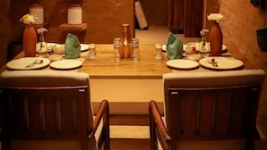 A dining area with a rustic stone wall, wooden chairs, a table set for two, and a hanging light fixture.