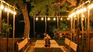An outdoor dining area with a table set for two, surrounded by wooden benches, under a large tree with string lights.