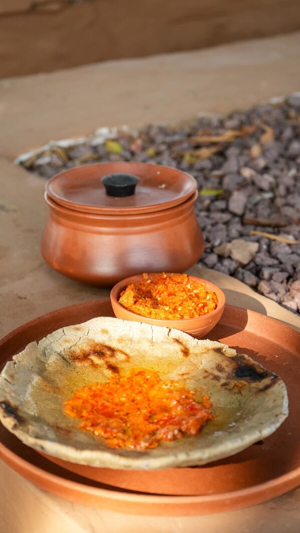 A flatbread with a red sauce on a clay plate, a small bowl of sauce, and a clay pot in the background.