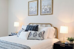 Ein ordentlich bezogenes Bett mit weißer Bettdecke, mehreren Kissen und einer blau-weißen Tagesdecke. Zwei Nachttische mit Lampen, ein kleiner Blumenstrauß in einer Vase und zwei gerahmte Bilder an der Wand sind zu sehen.