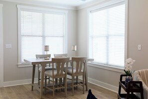 A dining area with a table and chairs, large windows with blinds, and a sofa with a vase. - "Sweet Home Mississippi" by OVR (Oxxford)